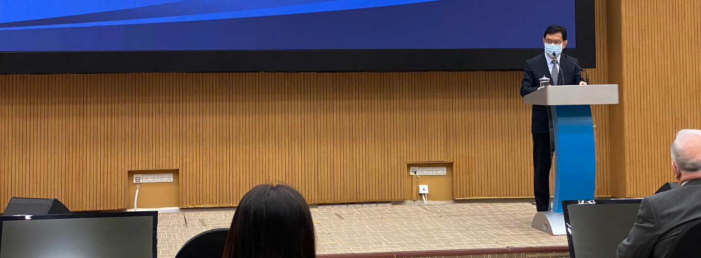 Person with mask at podium on stage with wooden paneling, monitor screen in foreground.